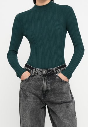 Personne portant un haut à manches longues ajusté, côtelé, vert foncé, rentré dans un jean noir ample et délavé, les mains dans les poches.