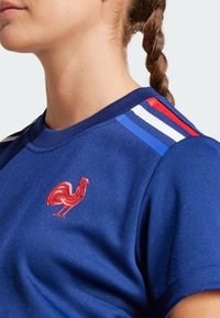 Blue athletic shirt featuring a red embroidered rooster logo and three vertical stripes in blue, white, and red on the shoulders.