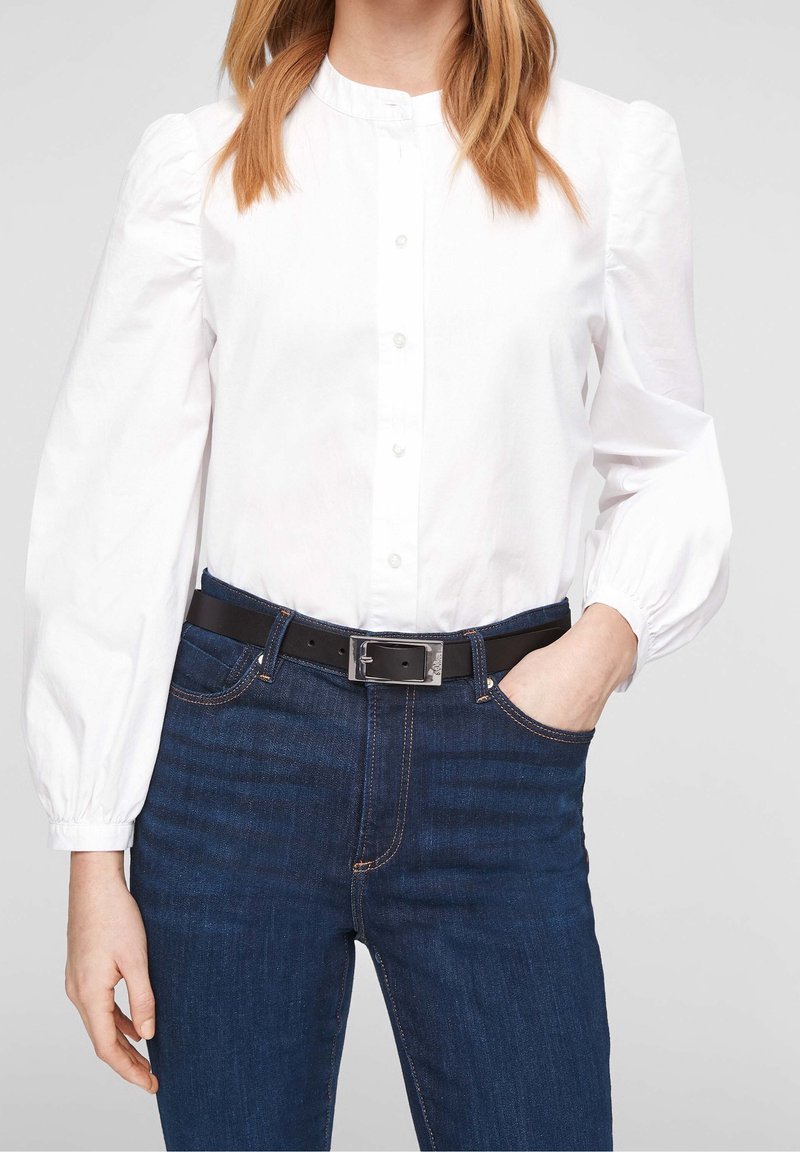 Femme portant un chemisier blanc à manches longues boutonné, rentré dans un jean bleu foncé avec une ceinture noire, une main dans la poche sur un fond uni.