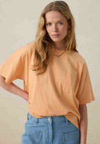 T-shirt à manches courtes, coupe décontractée en tissu doux pêche, avec une poche poitrine. Porté avec un short en denim taille haute bleu clair.