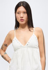 Νεαρή γυναίκα με μακριά σκούρα μαλλιά, που φοράει λευκό αμάνικο μπλουζάκι με ασημένια καρφιά και πολλά πολύχρωμα τατουάζ στα χέρια, κοιτάζει προς τα πλάγια.