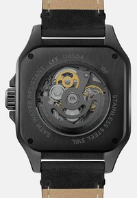 Schwarze Edelstahl-Uhr mit quadratischem Gehäuse, transparentem Rücken, der komplizierte Zahnräder zeigt, und einem schwarzen Lederarmband mit beigen Akzenten.