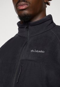 Czarna polarowa kurtka z wysokim kołnierzem, zamkiem z przodu i boczną kieszenią. Posiada małe białe logo Columbia na piersi. Miękka textura.