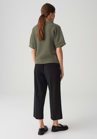 Top corto a maniche corte verde oliva con texture abbinato a pantaloni neri a gamba larga e mocassini neri, visto da dietro.