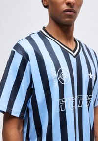 Άνδρας που φοράει αθλητική φανέλα με κοντά μανίκια και V-λαιμόκοψη, με εναλλασσόμενες κάθετες ρίγες ανοιχτού και σκούρου μπλε χρώματος και λευκά γραφικά λογότυπα στο στήθος.