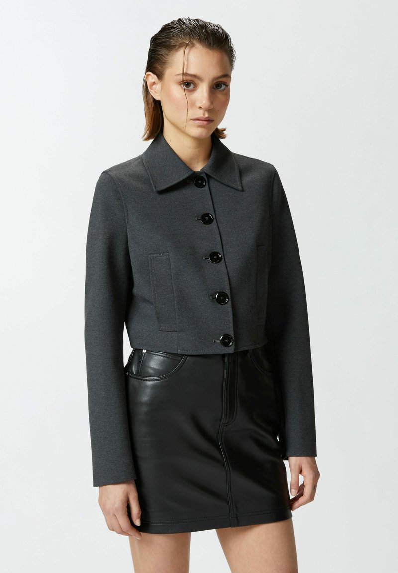 Giacca grigia corta realizzata in tessuto testurizzato, con colletto a punta, sei bottoni neri e due tasche frontali, abbinata a una gonna in pelle nera.