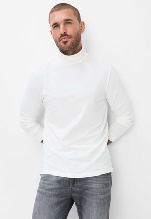 Man met kort haar en baard, draagt een witte coltrui en grijze spijkerbroek, staand tegen een witte achtergrond.