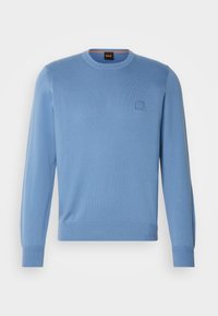 Pull tricoté bleu clair avec un col rond, des manches longues et un petit patch logo carré sur la poitrine gauche. Tissu texturé.