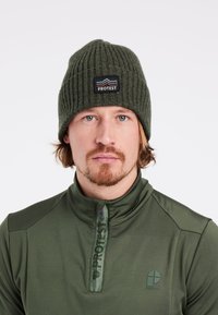 Grüne Strickmütze mit gerippter Textur, ausgestattet mit einem schwarzen Aufnäher mit der Aufschrift 'PROTEST' und einem Berggrafik. Getragen mit einem grünen Zip-Up-Oberteil.
