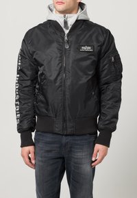 Sort bomberjakke lavet af nylon, med en frontlynlås, ribbede manchetter og en grå hætte under. "Alpha Industries" logo på ærmet.