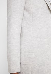 Svetle šedý blazer s texturovaným herringbonovým vzorem, strukturovanými rameny a jednou náprsní kapsou. Tkanina vypadá měkce a tkaně.