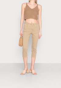 Beige zkrácený top s pleteným designem, doplněný o světlé béžové capri kalhoty a světle růžové muly na blokovém podpatku. Drží tkanou tašku.