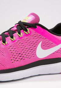Scarpa da corsa Nike di un brillante colore rosa con lacci neri, occhielli verdi, logo swoosh bianco e suola bianca, su uno sfondo semplice.