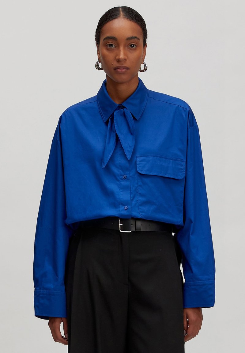 Blauw oversized overhemd met een stropdas, knoopsluiting aan de voorkant, lange mouwen en een borstzak; gladde textuur en ruime pasvorm.