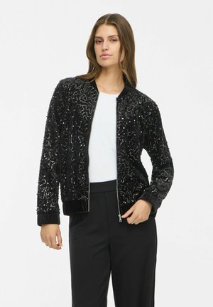 Mujer con chaqueta bomber negra con lentejuelas sobre camiseta blanca y pantalones negros, de pie frente a un fondo claro y liso.