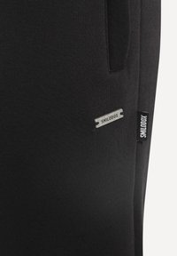 Sorte sportbukser med ribbet tekstur. Har en sølvfarvet metaltag med mærket "SMILODOX" og en stoffetiket på siden.