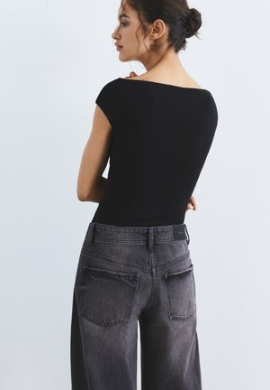 Femme aux cheveux foncés attachés, portant un haut noir ajusté et un jean noir délavé taille haute, tournée légèrement de profil sur un fond uni.