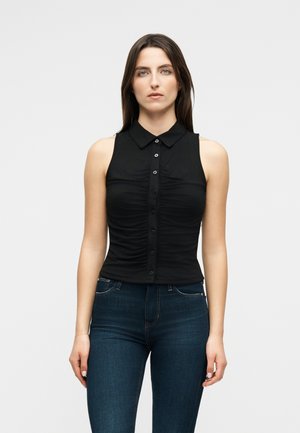 Naine kannab varrukateta musta triiksärgiga, millel on krae ja nööbid, tumedaid siniseid kitsaid teksapükse, seistes tühja heleda tausta ees.