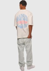 Creme oversized t-shirt med blå og lyserød grafisk tekst på ryggen, parret med lysegrå baggy jeans og hvide sneakers.