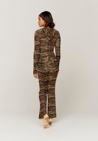 Tettsittende jumpsuit i teksturert stoff med et brunt og gull sikksakkmønster. Har lange ermer og hette, kombinert med beige hæler.