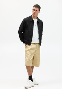 Leichte schwarze Jacke mit Reißverschluss, kombiniert mit einem weißen T-Shirt und beigen knielangen Shorts. Weiße Sneakers runden das Outfit ab.