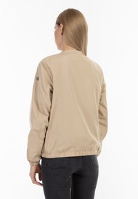 Beige bomber bunda vyrobená z hladkého, lehkého materiálu, s elastickými manžetami a přiléhavým pasem. Má kulatý výstřih a logo na rameni.