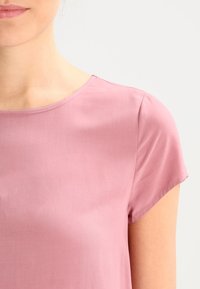 Lichtroze blouse met korte mouwen, ronde halslijn en een gladde, zachte stof. Naaddetails zijn zichtbaar op de schouders.