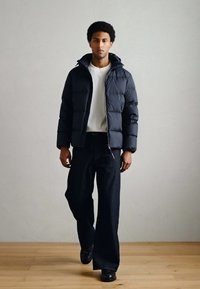 Casaco acolchoado em azul-marinho escuro, com fecho de correr, capuz e textura acolchoada. Combinado com jeans pretos de perna larga e sapatos pretos.