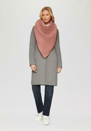 Manteau en laine gris avec un châle texturé en forme de triangle de couleur rose drapé autour du cou. Associé à un jean noir et des baskets blanches.