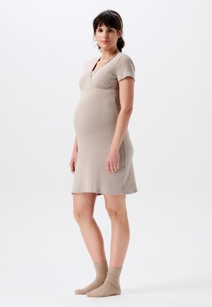Zwangere vrouw die staat, gekleed in een beige jurk met korte mouwen en bijpassende sokken, tegen een effen witte achtergrond.