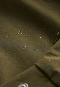 Tissu olive vert résistant à l'eau avec une texture lisse, présentant des gouttes d'eau à la surface et un détail de bouton métallique.
