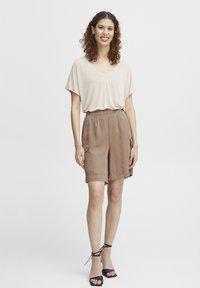 T-shirt beige à col en V associé à un short marron ample avec des poches latérales. Des sandales à talons strappy noires complètent la tenue.