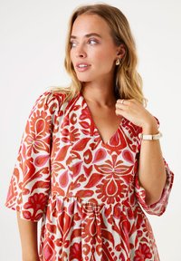 Vestido de colores rojo y rosa con un escote en V y mangas amplias, que presenta diseños florales sobre un fondo claro. Acentuado con una pulsera.
