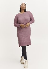 Abito rosa lavorato a maglia con maniche lunghe, polsini a costine, spacchi laterali e una vestibilità morbida. Abbinato a collant neri e scarpe da ginnastica bianche chunky.