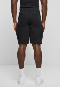 Shorts cargo noirs à coupe décontractée, dotés de poches latérales, d'une taille plate et d'une texture en coton lisse. Baskets et chaussettes blanches visibles.