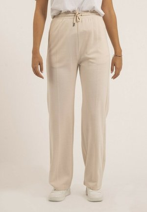 Femme portant un pantalon beige à jambes larges avec une taille à cordon de serrage, des baskets blanches, debout devant un fond clair uni.