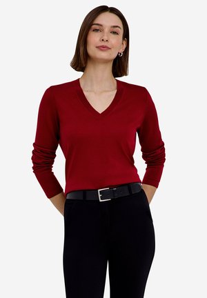 Mujer con el pelo corto y castaño, vistiendo un suéter rojo con cuello en V, pantalones negros y un cinturón negro, de pie con las manos en los bolsillos.