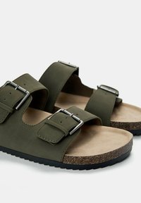 Due sandali verde oliva con due cinturini regolabili, fibbie argentate e suola in sughero. Il fondino è realizzato in un materiale liscio di colore beige.