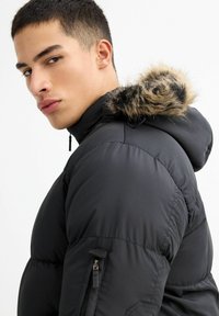 Chaqueta acolchada negra con capucha forrada de piel sintética, que cuenta con un bolsillo lateral con cremallera y una textura acolchada. Diseño ajustado, ideal para climas fríos.