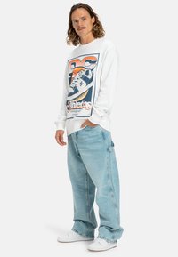 Witte sweatshirt met een grafisch ontwerp van een sneaker en het woord "Athletics", gecombineerd met losse, lichtblauwe jeans en witte sneakers.