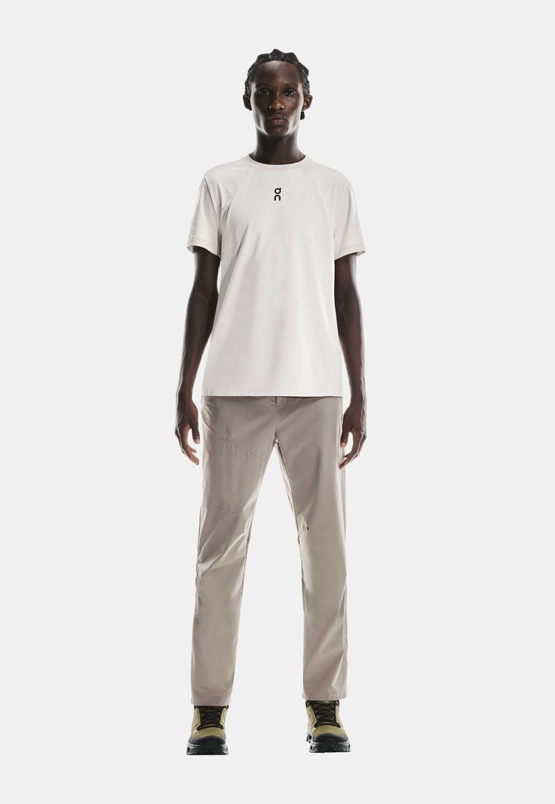 T-shirt grigio chiaro a maniche corte con piccolo logo, abbinato a pantaloni cargo beige e scarponi da trekking neri con dettagli gialli.