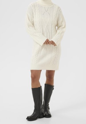 Femme portant une robe-pull blanche en maille torsadée à col roulé avec des bottes en cuir noires montant jusqu'aux genoux, debout sur un fond uni.