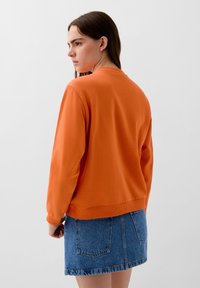 Felpa arancione con collo e polsini a costine, abbinata a una gonna di jeans. Il tessuto sembra morbido e ha una texture liscia.