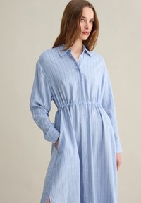 Abito bluu chiaro con bottoni, a righe verticali, maniche lunghe, vita arricciata e tasche laterali. Tessuto morbido e testurizzato.