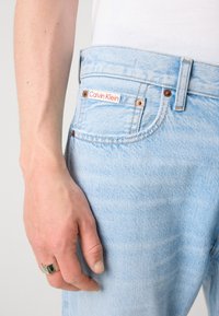 Svijetloplave traperice s izblijedjelom teksturom, imaju crvenu i bijelu Calvin Klein oznaku blizu struka i brončane metalne detalje.