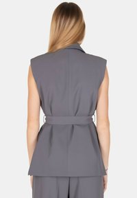 Blazer grigio senza maniche con un design strutturato, caratterizzato da una cintura in vita e un colletto spigoloso. Tessuto liscio con una leggera lucentezza.