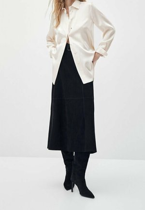Personne portant une chemise crème à boutons, une jupe midi noire avec une ceinture, et des bottes noires à talons montant jusqu'aux genoux, debout les mains dans les poches.