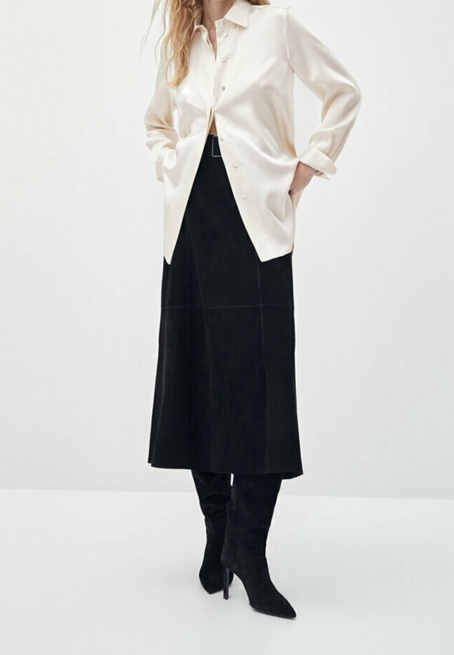 Personne portant une chemise crème à boutons, une jupe midi noire avec une ceinture, et des bottes noires à talons montant jusqu'aux genoux, debout les mains dans les poches.