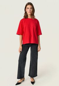 Red cotton oversized t-shirt with short sleeves, featuring a small textured logo pocket. Paired with black wide-leg jeans and black heels.