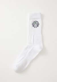 Bílá bavlněná ponožka s žebrovaným lemem, na které je kulaté logo Lacoste Tennis Club v námořnické modři, včetně zkřížených raket a vavřínových akcentů.